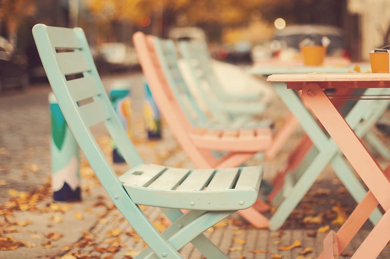Row of pastel blue and pink outdoor cafe chairs and tables on a brick sidewalk with blurred background.