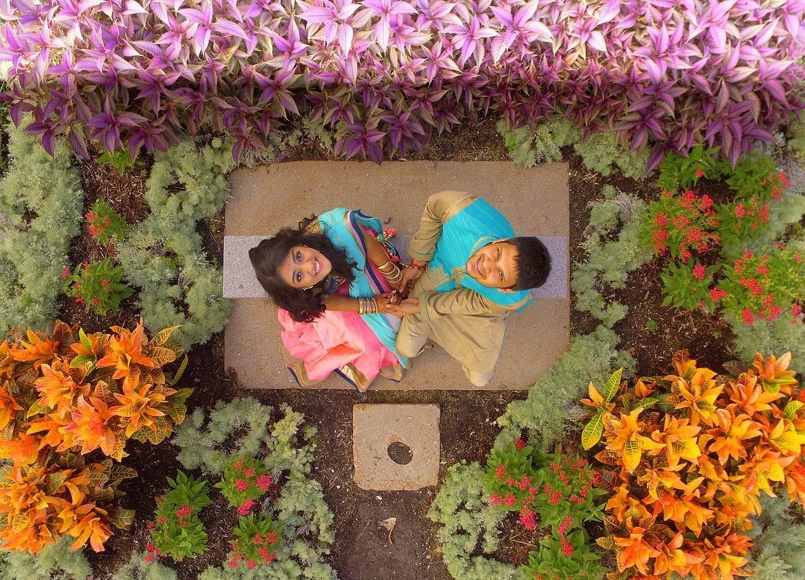 Couple holding hands, seated on a stone bench surrounded by colorful flowers. Overhead view.