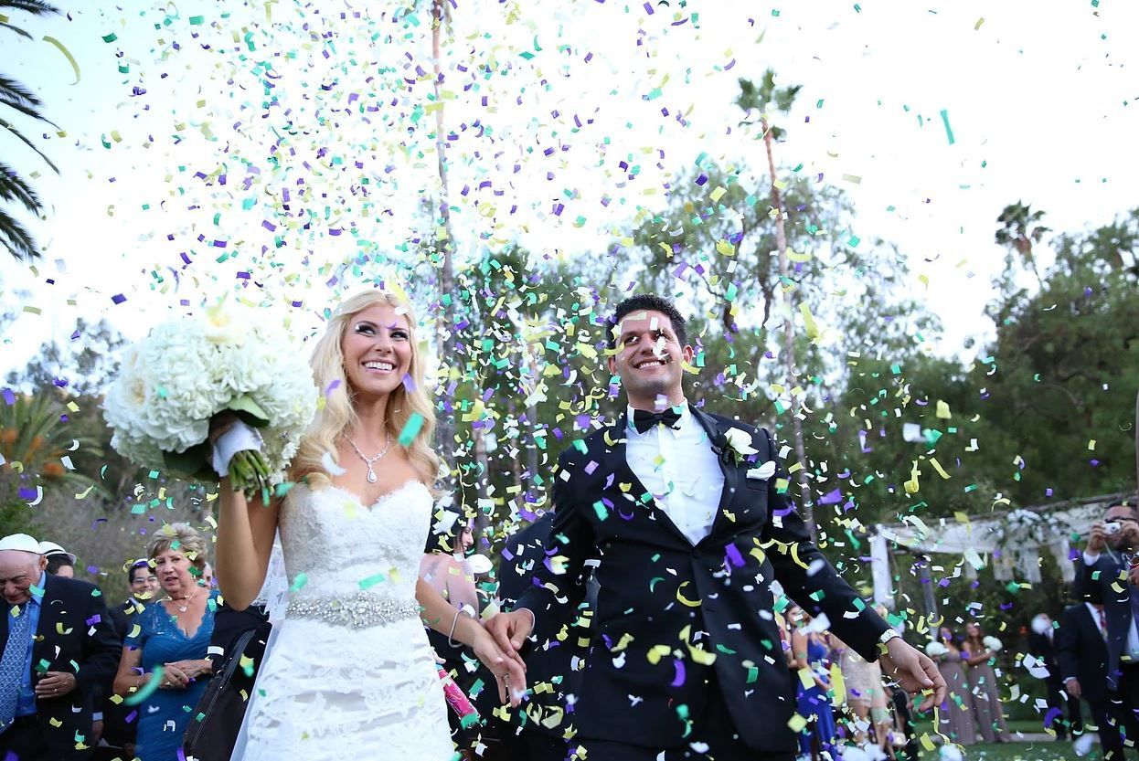 Bride and groom exiting wedding ceremony, confetti falling. Bride smiles, holds bouquet. Groom smiles, wears tuxedo.