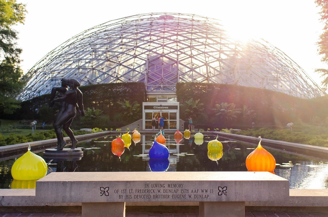 Dome of glass building with a sculpture and colorful glass art on a pond, illuminated by sunlight.