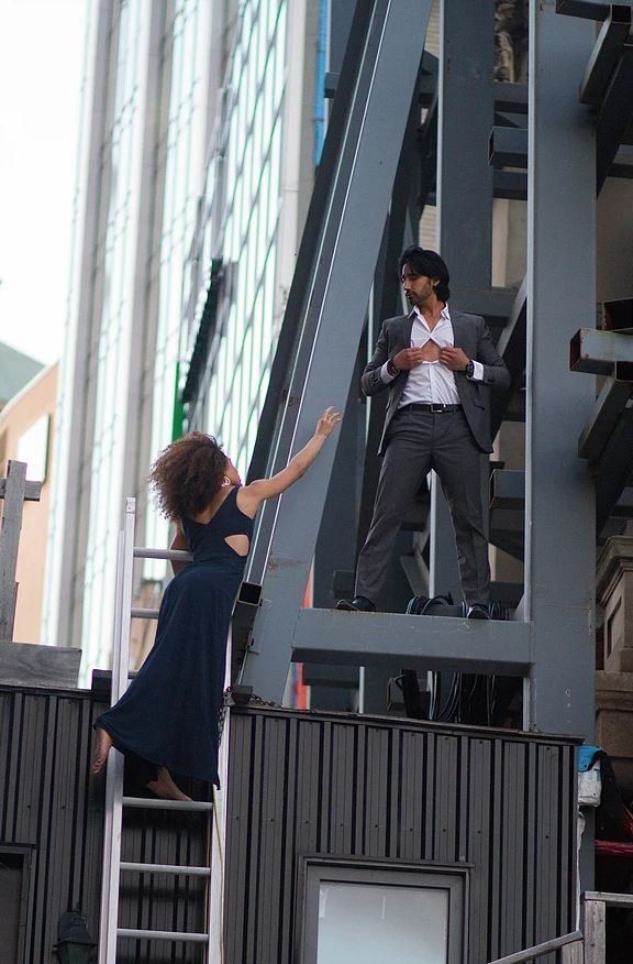 Woman in blue dress reaching towards man in a suit standing on a metal structure in a city setting.
