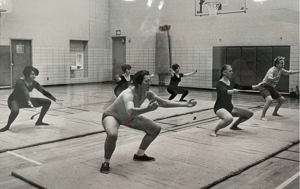 People in a gym doing synchronized squats on mats, with a basketball hoop in the background.
