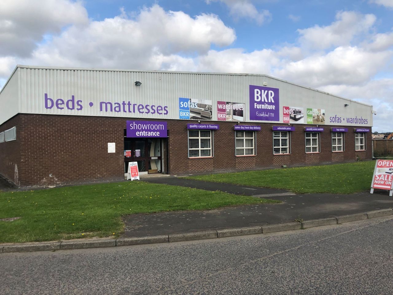 BKR Beds & Mattresses store exterior, brick building, purple logo, sign, windows, green grass, cloudy sky.