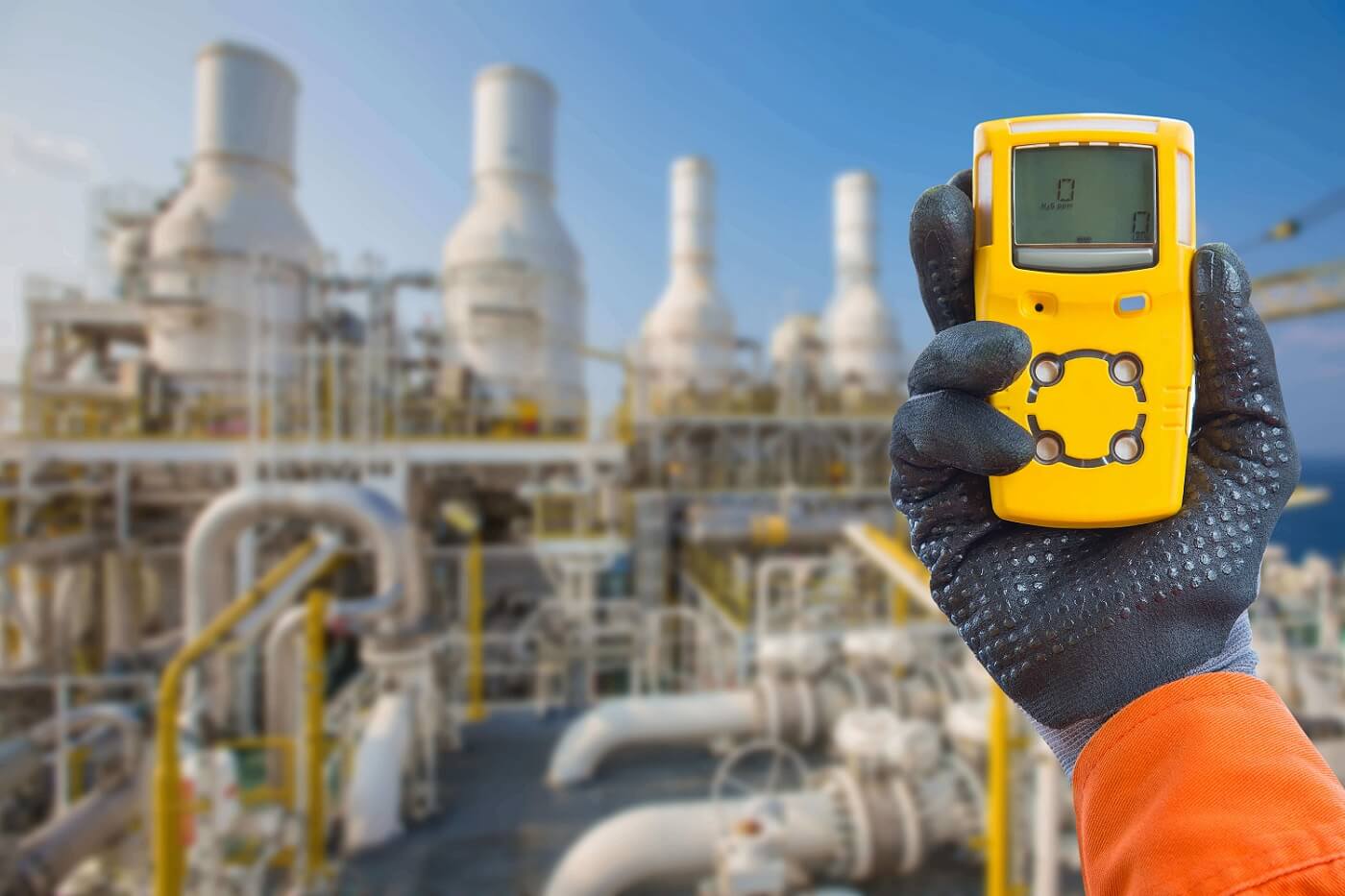 Hand met handschoen die een gele gasdetector vasthoudt in een industriële installatie met pijpleidingen en torens.