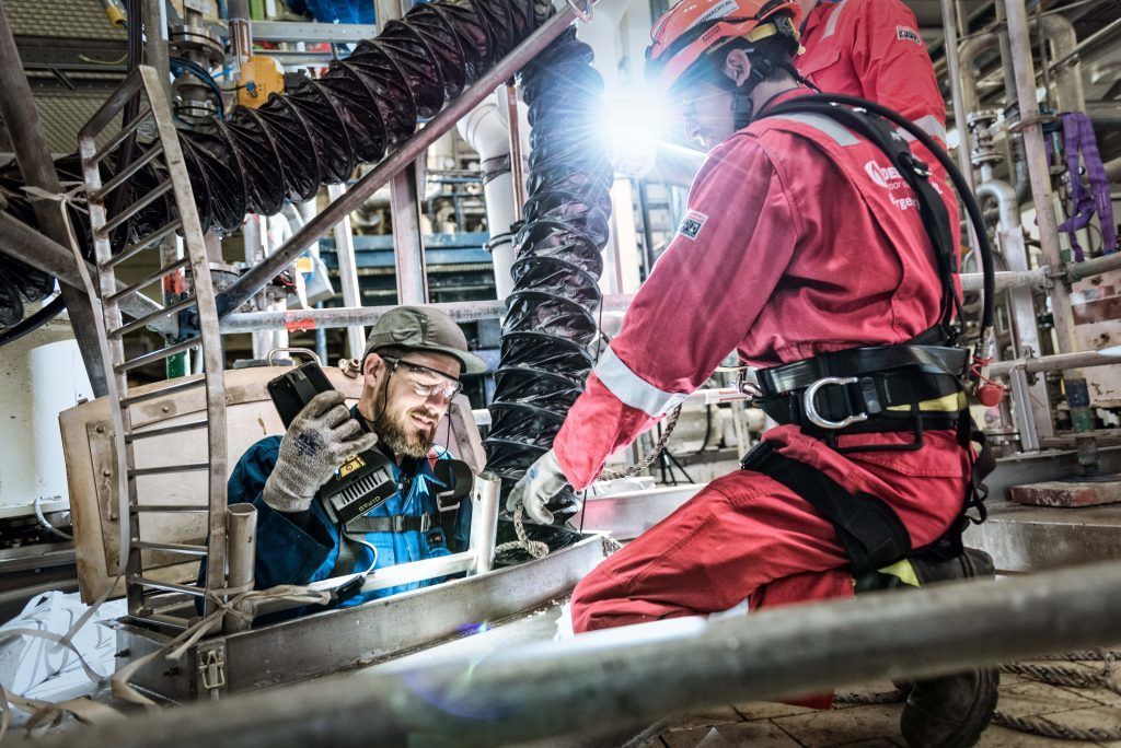 Werknemers in rode veiligheidskleding inspecteren industriële leidingen in een fabriek; een van hen hurkt en een ander staat.