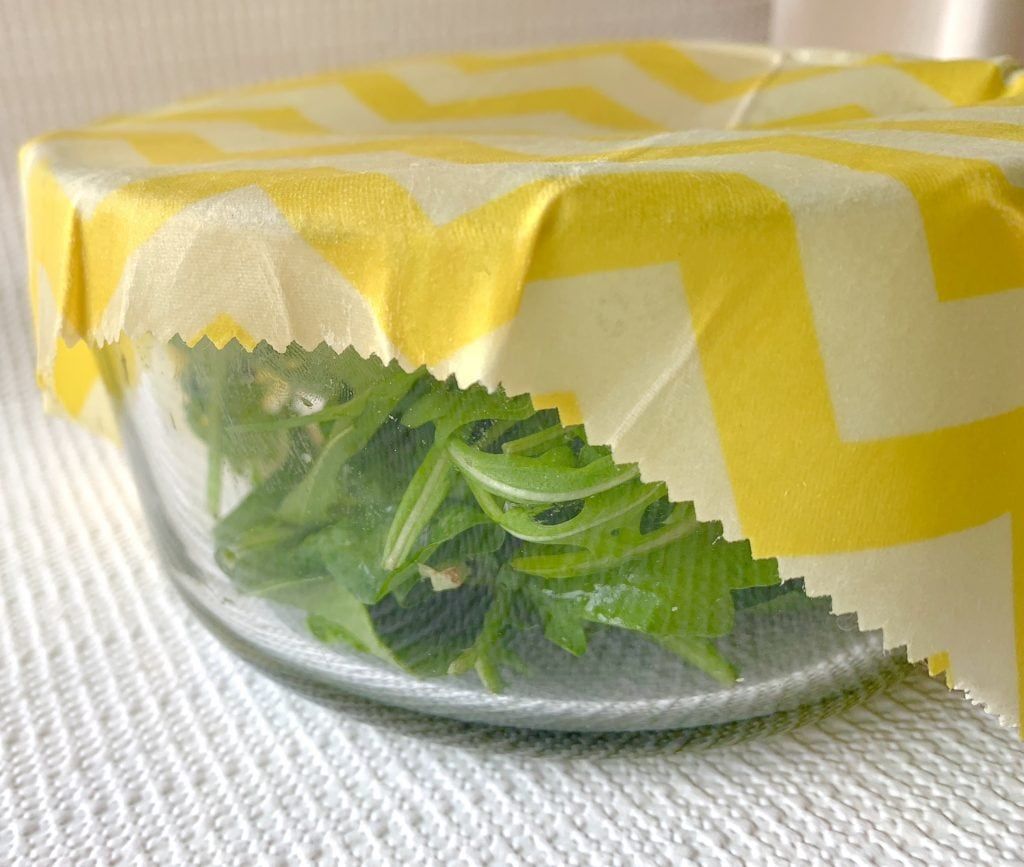 A glass bowl covered with yellow-patterned cloth, with leafy greens visible inside.