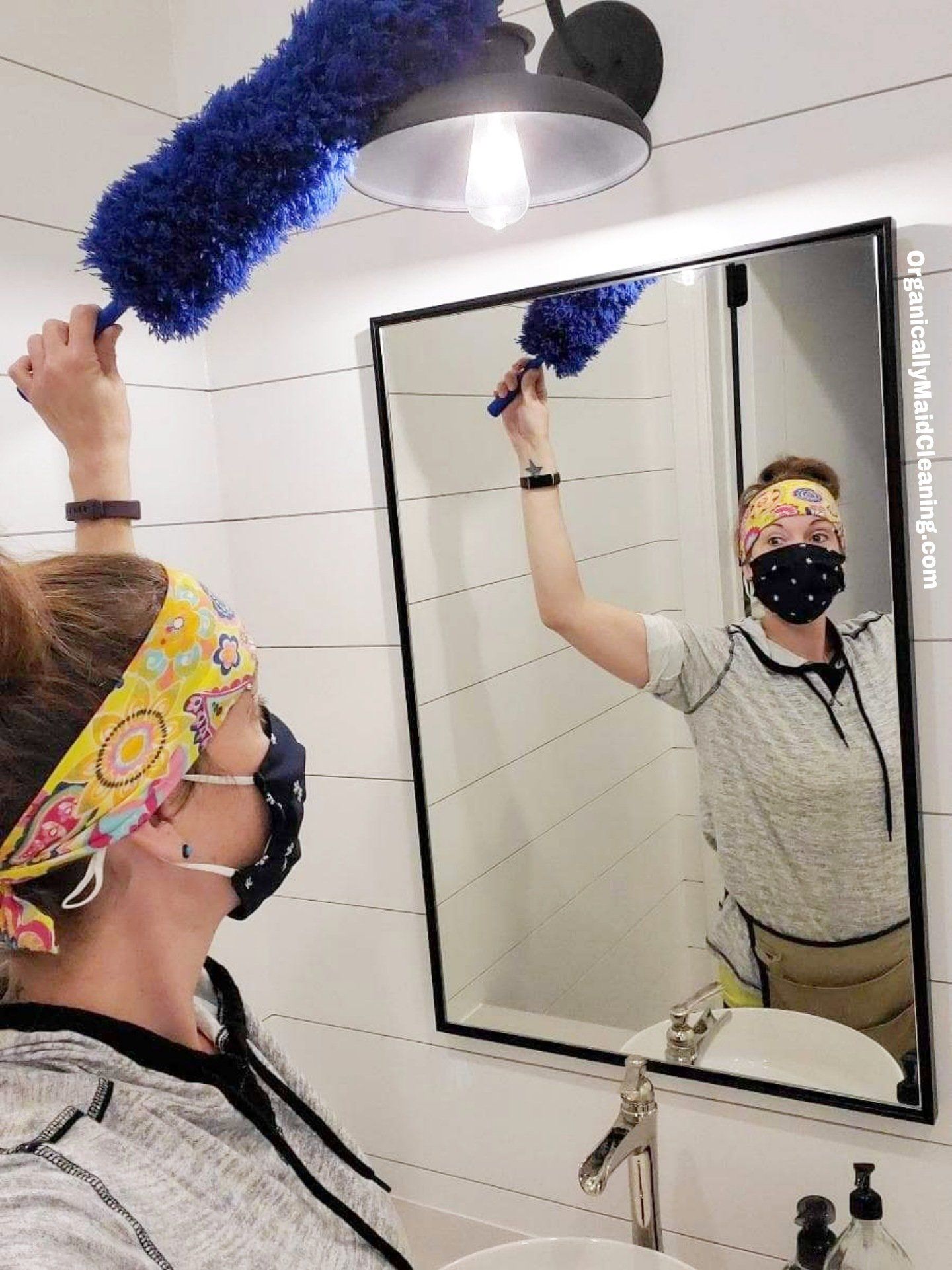 A person wearing a patterned headband and a face mask uses a blue duster to clean a light fixture above a bathroom mirror.