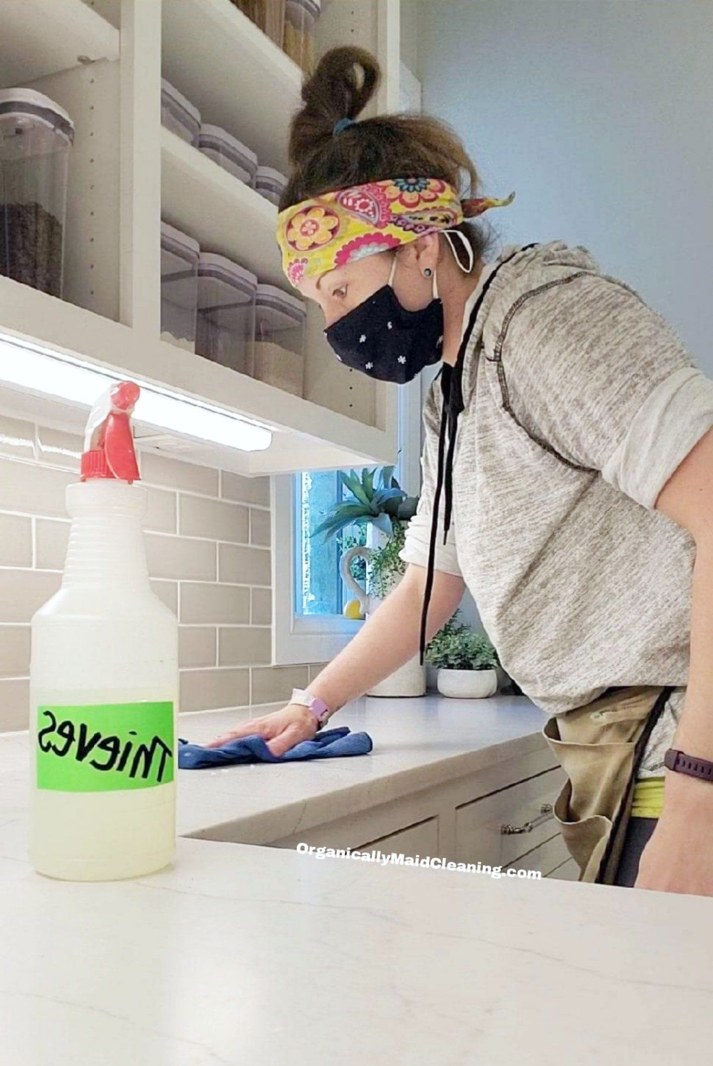 A person wearing a patterned headband and a black mask wipes a kitchen counter next to a bottle labeled 