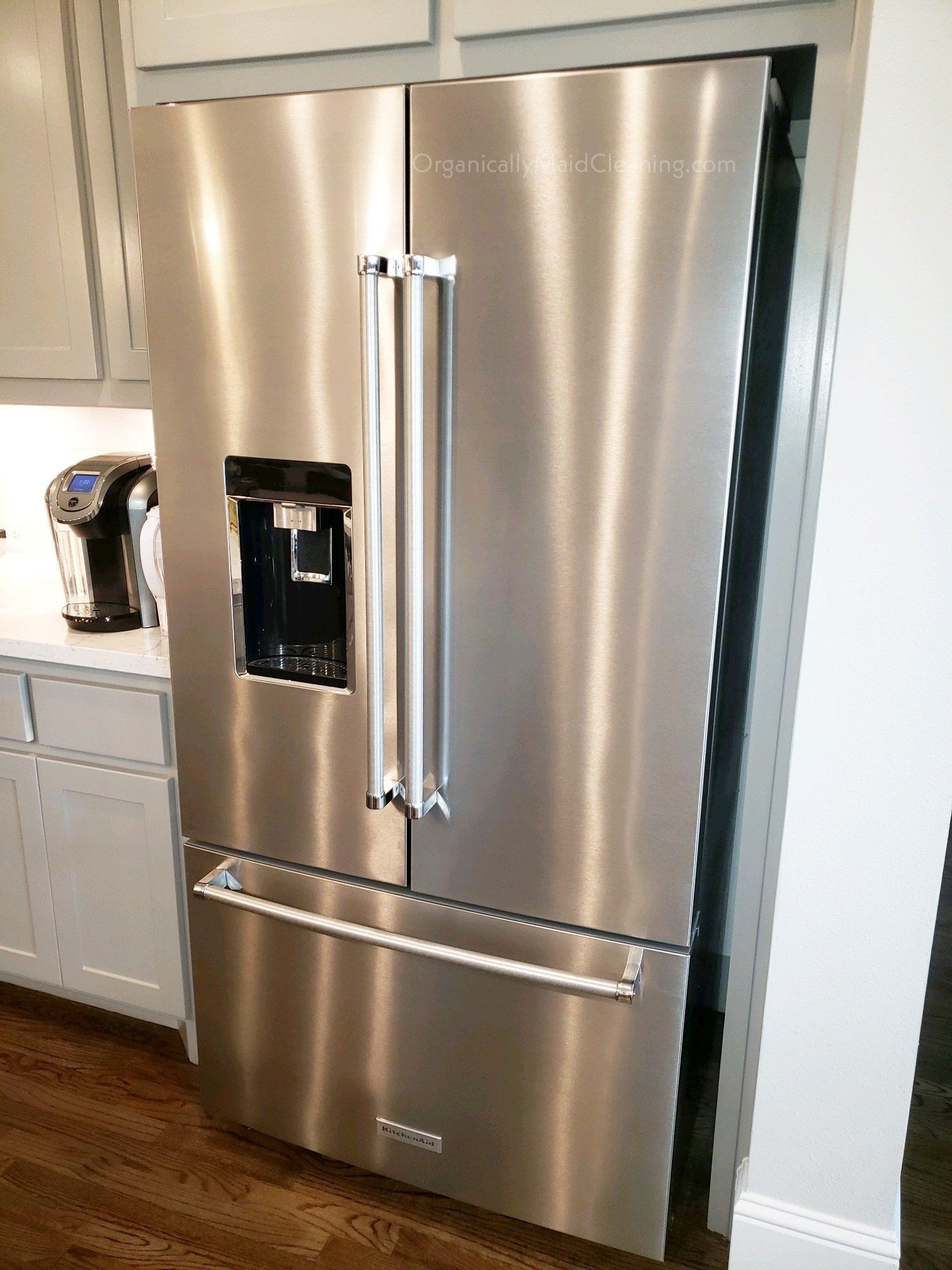 A stainless steel French door refrigerator with an external water dispenser, installed in a kitchen with gray cabinets.