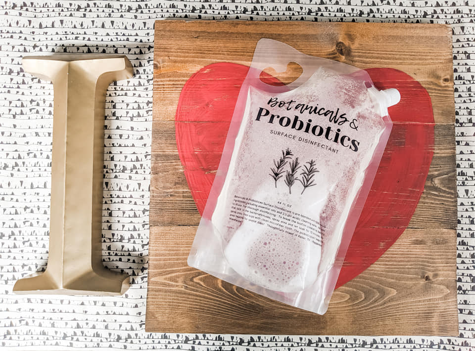 A pouch of Botanicals & Probiotics sits on a wooden board with a painted red heart, next to a large letter “I”.