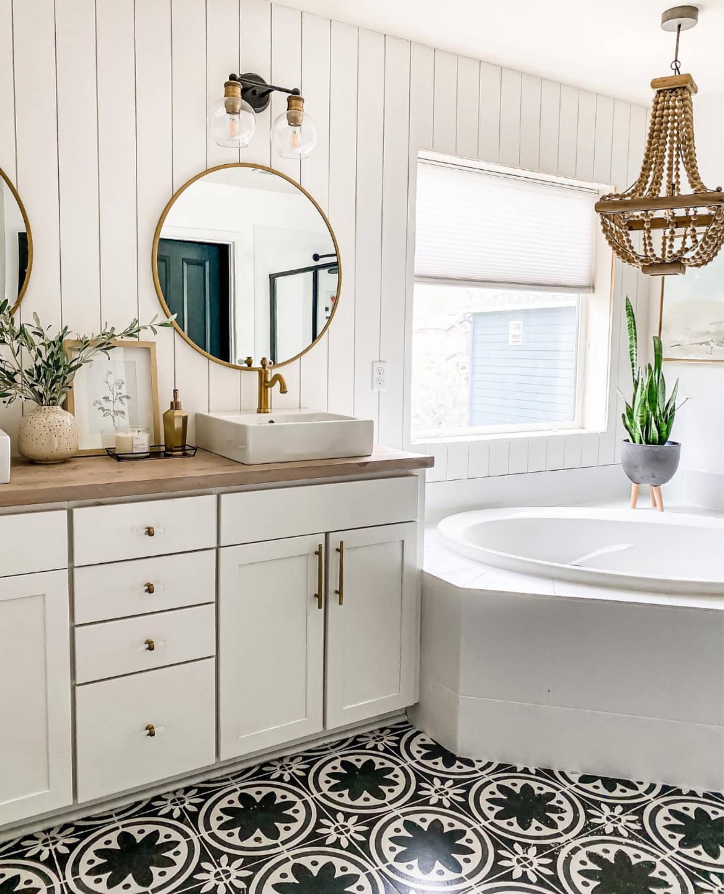 A bright, modern bathroom with a white vanity, circular mirror, beadboard wall, patterned tile floor, and a soaking tub.