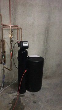 A black water softener is sitting in a basement next to copper pipes.