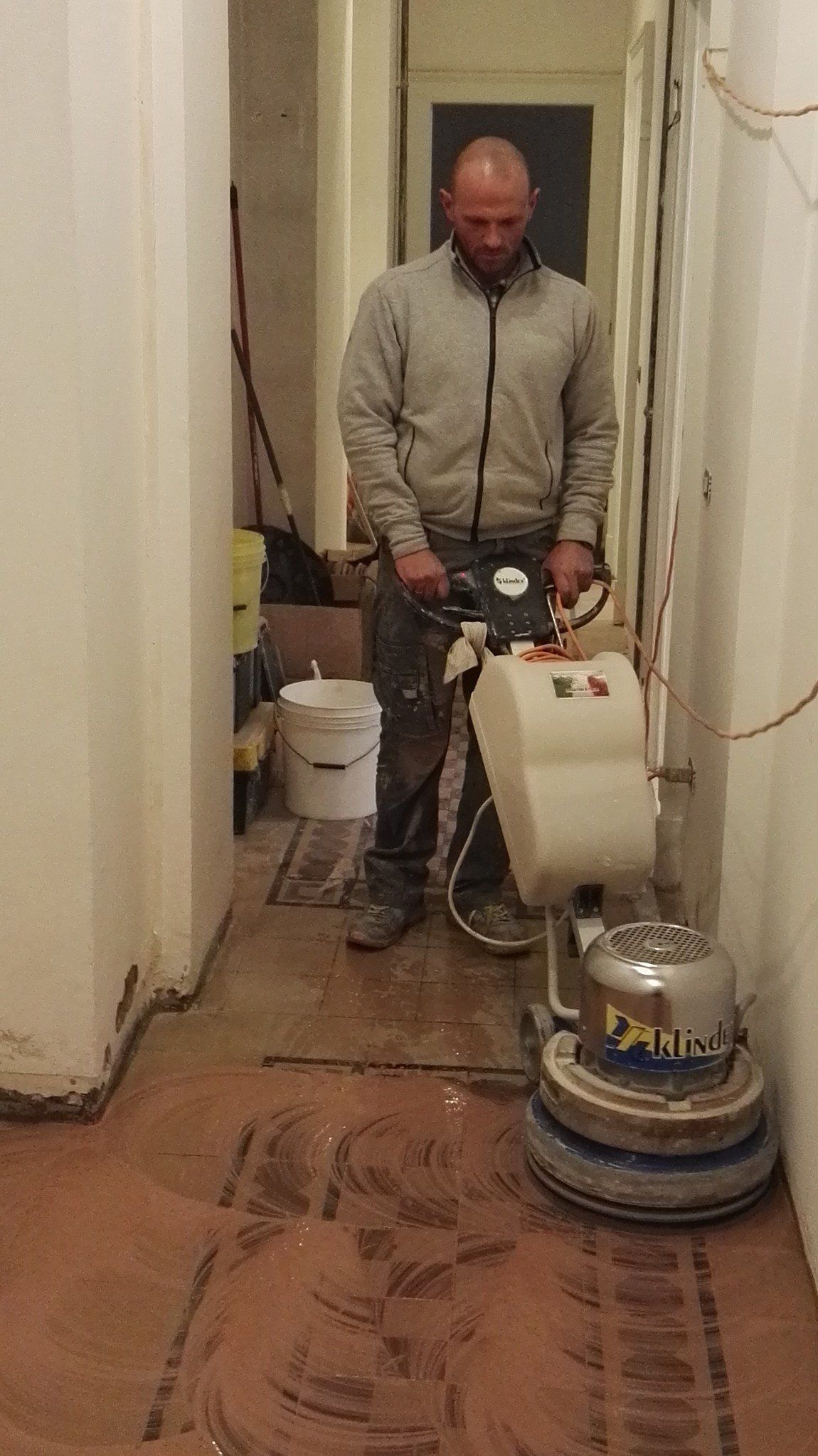 Man sanding a wooden floor in a narrow hallway. He wears a grey jacket and holds a floor sander.