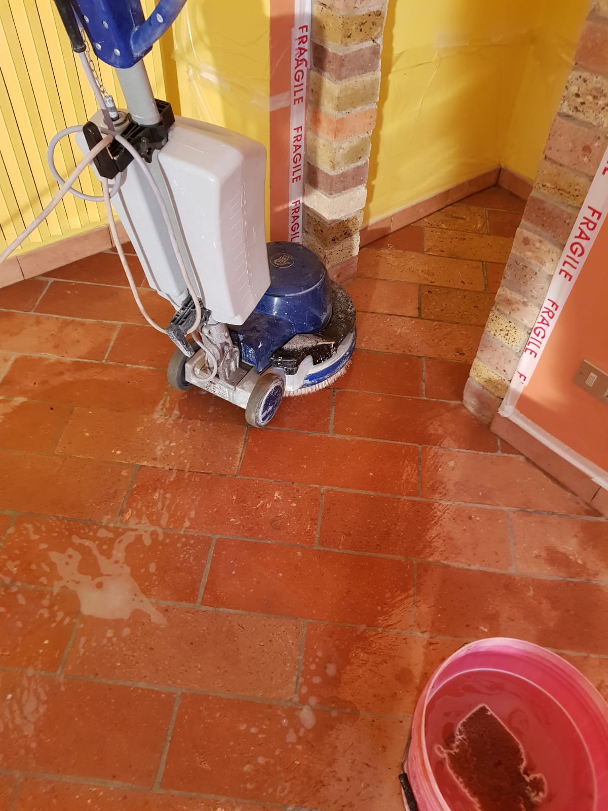 A floor cleaning machine scrubbing terracotta tiles in a corner of a room.