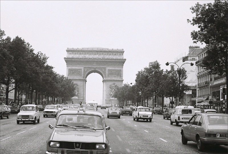 A black and white photo of cars driving down a street with a triumphal arch in the background