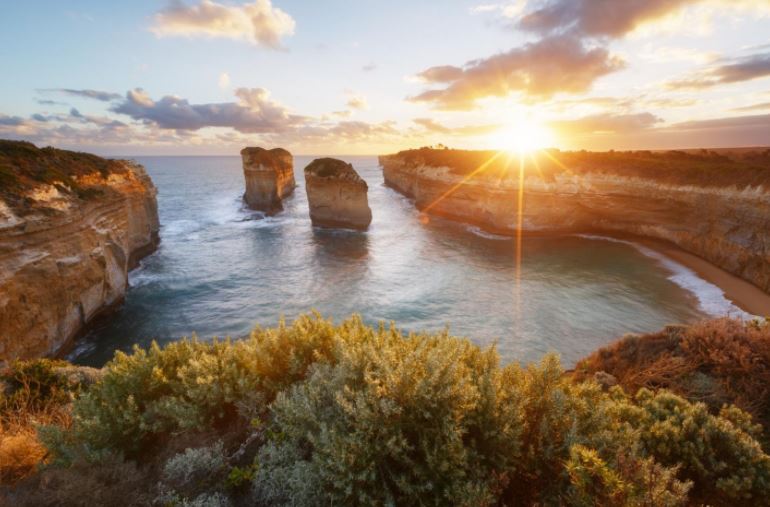 The sun is setting over a body of water with rocks in the foreground.