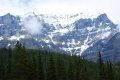 A snowy mountain with trees in the foreground and a cloudy sky in the background.