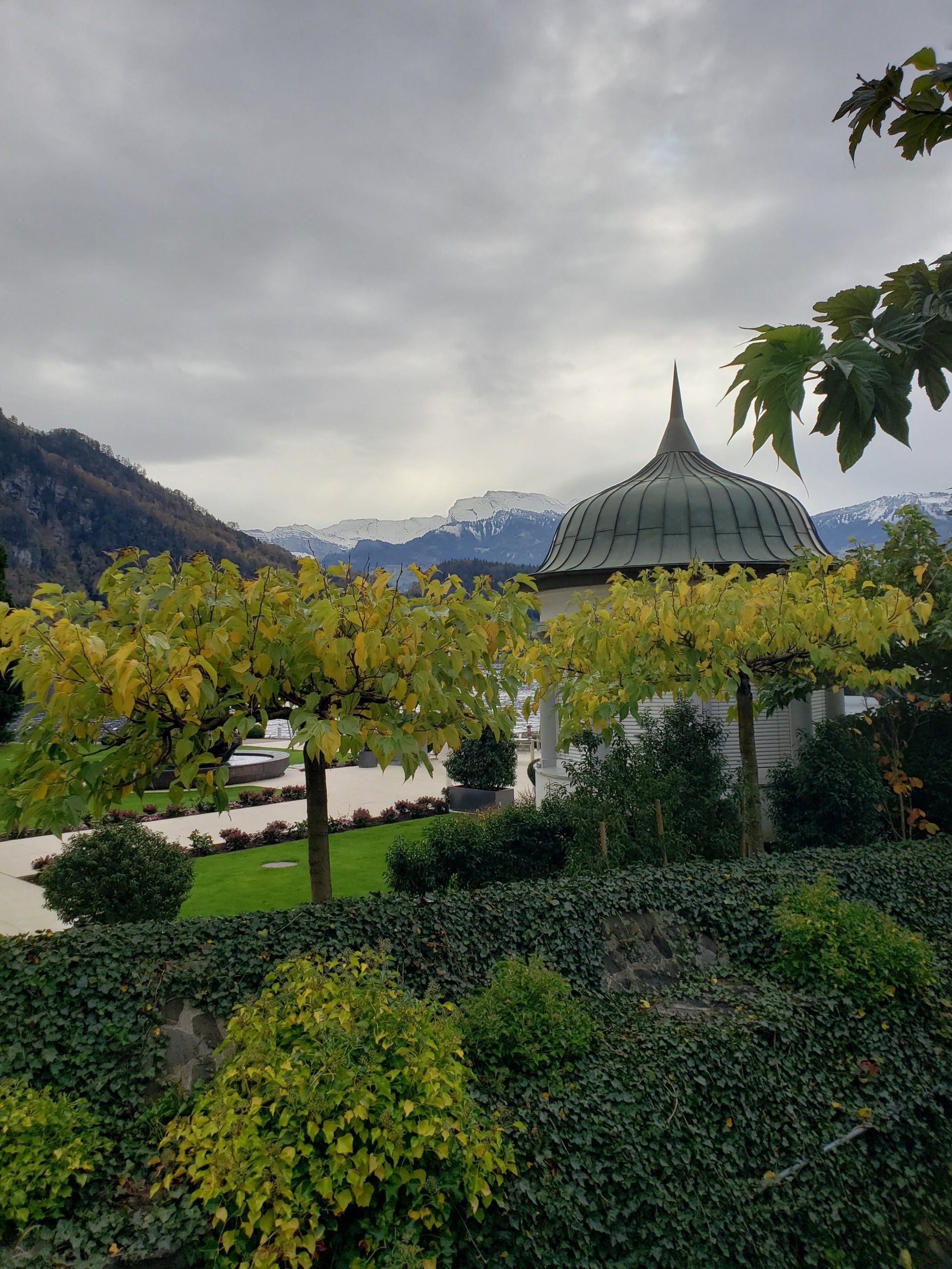 A gazebo is surrounded by trees and bushes in a park with mountains in the background.