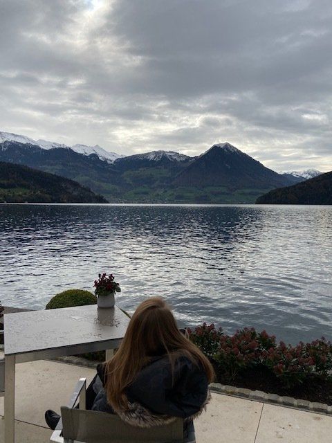 A woman sits at a table overlooking a lake with mountains in the background
