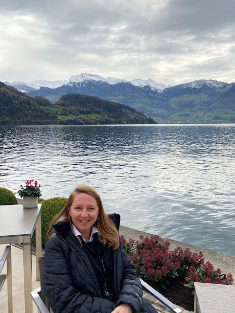 A woman is sitting in front of a lake with mountains in the background.
