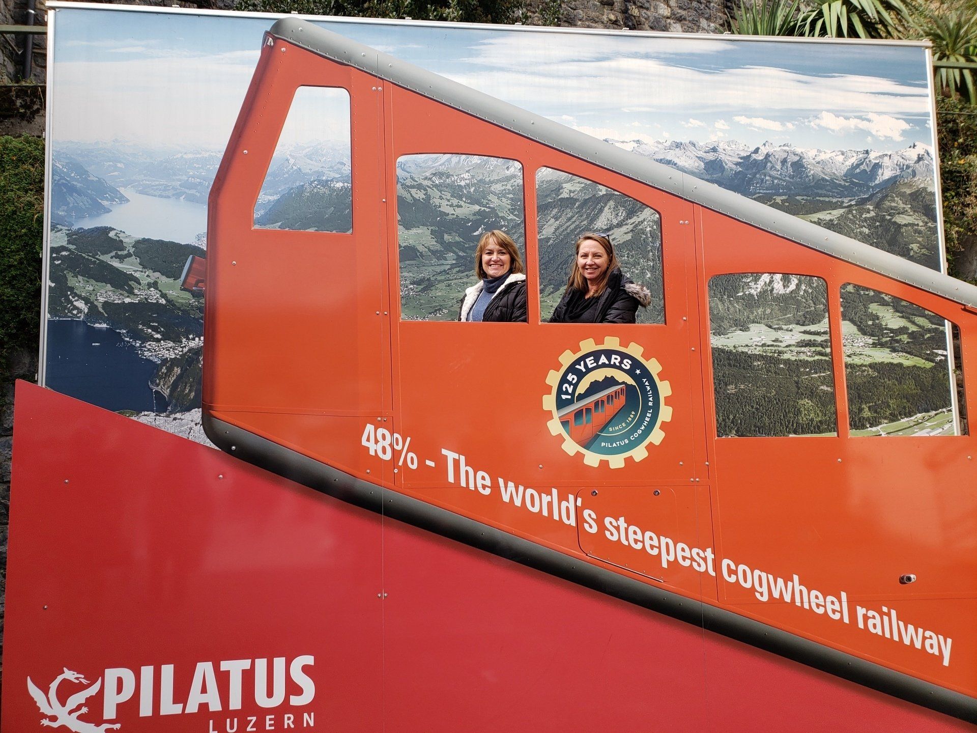 Two women are posing for a picture in front of an advertisement for the world 's steepest cogwheel railway