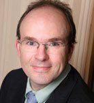 A headshot of a person wearing glasses, a light green collared shirt, and a dark blazer against a plain beige background.