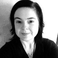 A black and white close-up portrait of a person with dark hair, wearing a dark v-neck top and a small necklace.