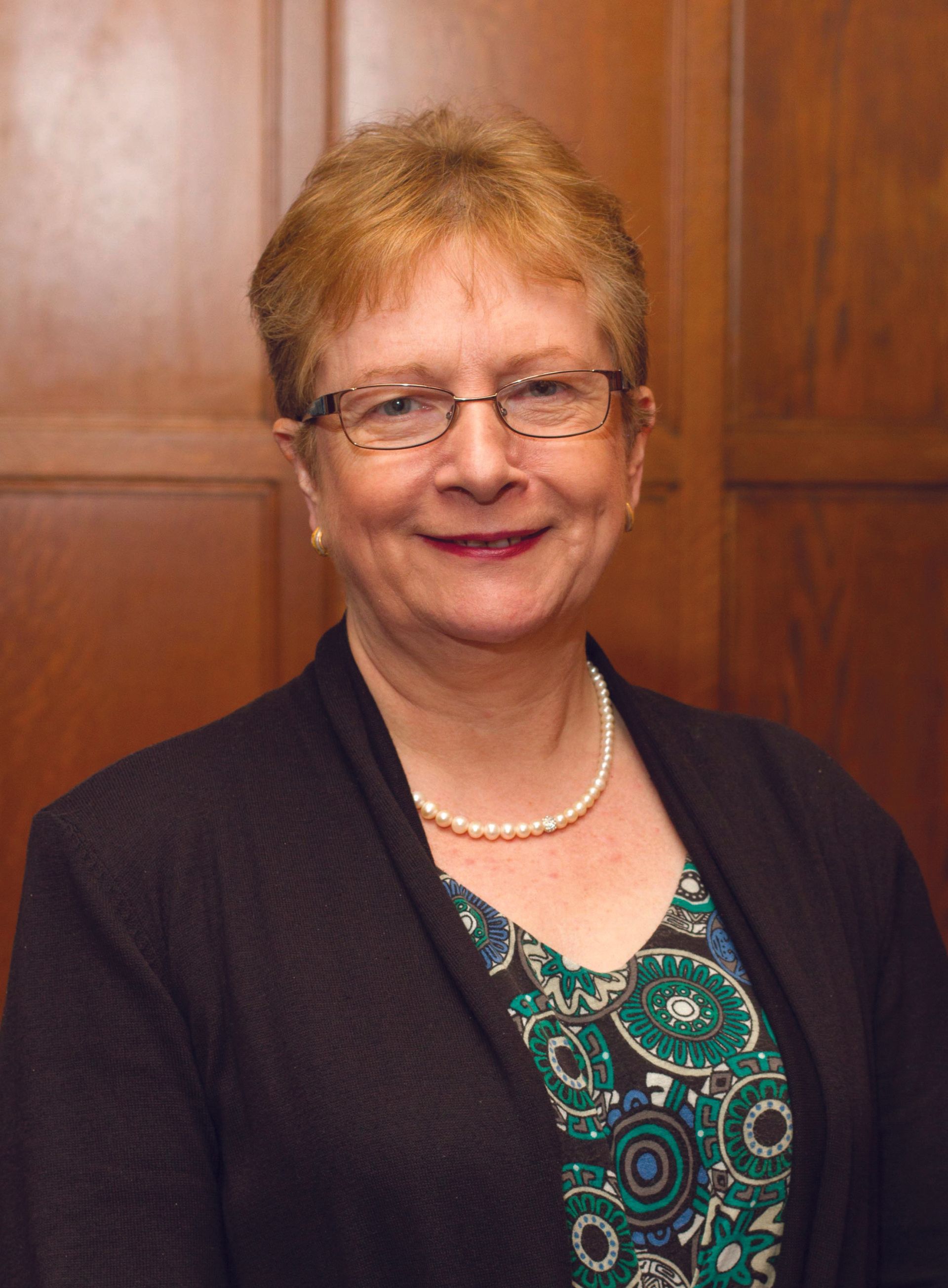 A person with short, reddish hair and glasses smiles while wearing a pearl necklace and a patterned green top.