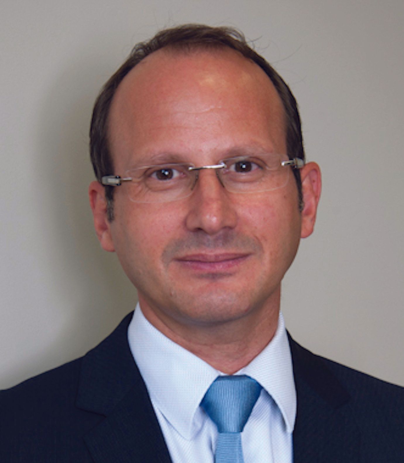A person wearing a suit, white shirt, and light blue tie with rimless glasses, captured in a headshot against a light wall.