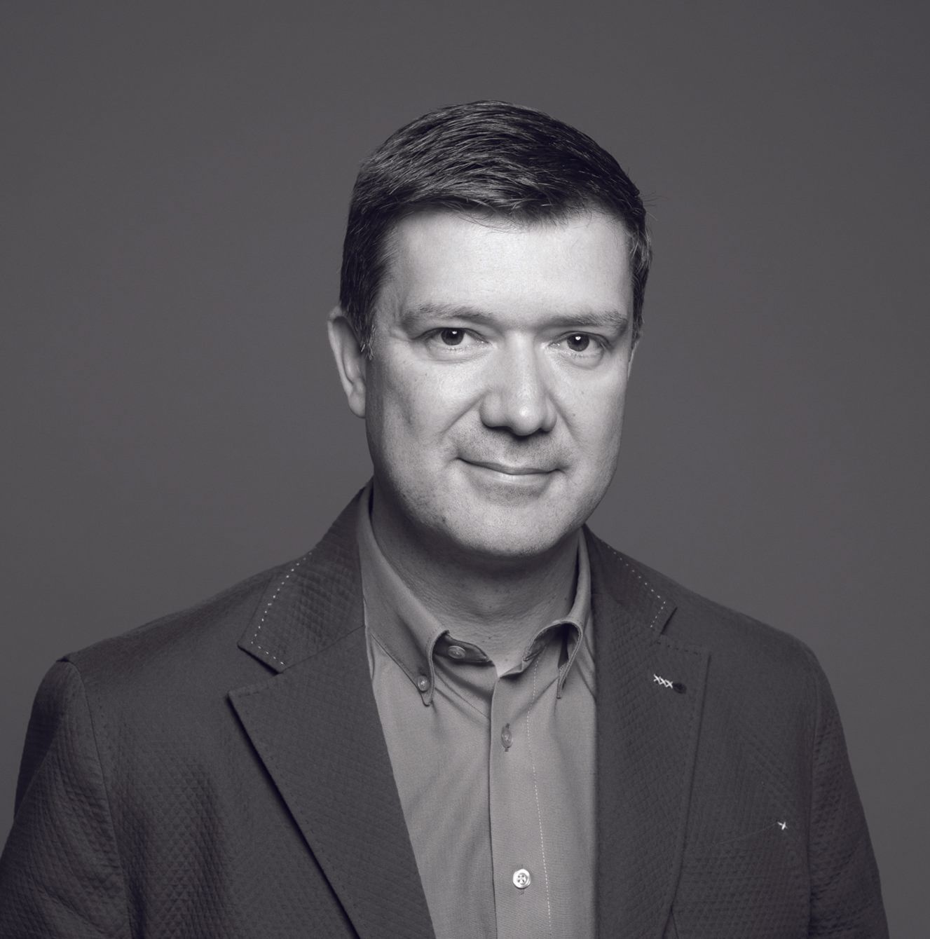 Headshot of a person wearing a button-down shirt and blazer against a plain gray background.