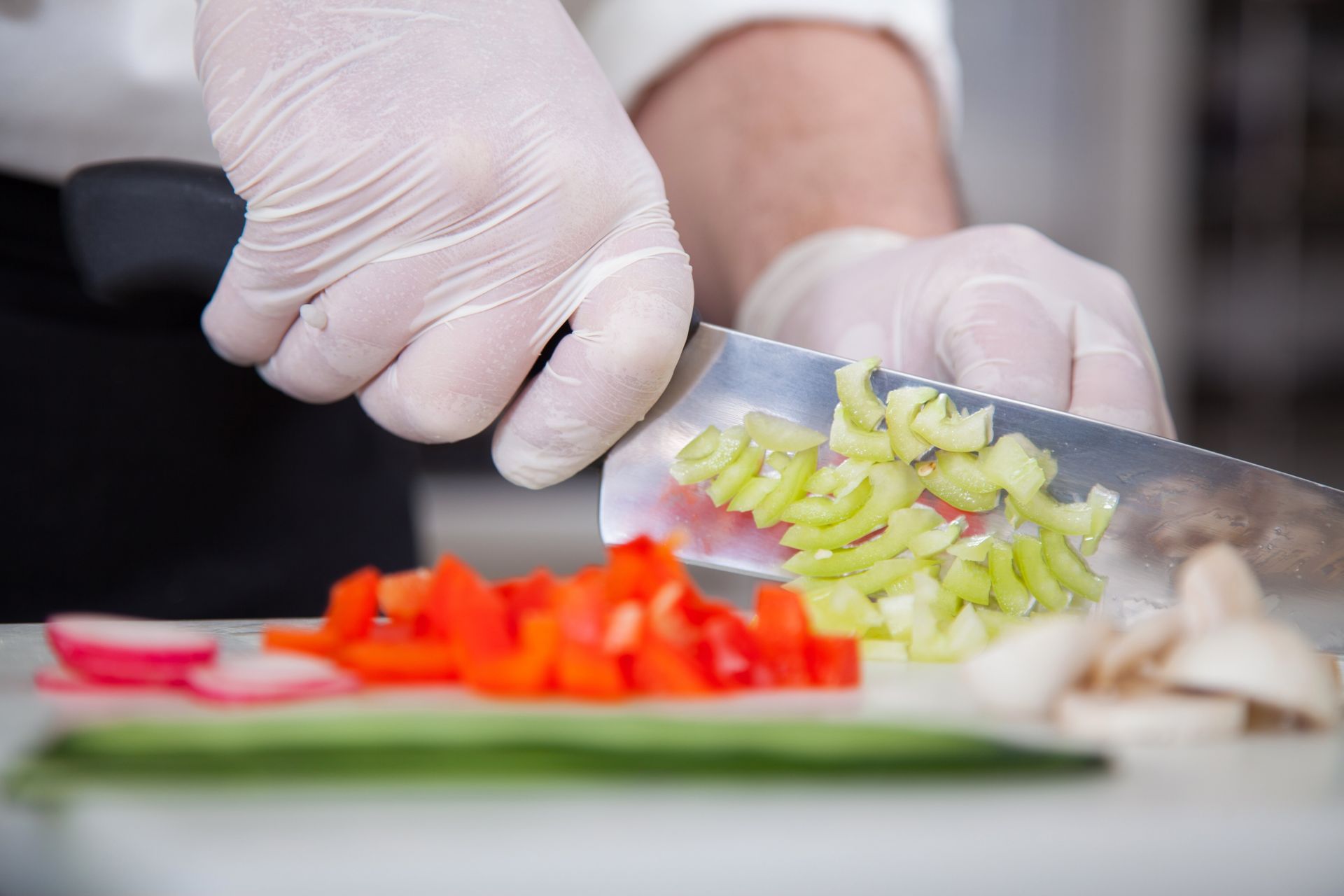 Ein Koch mit Handschuhen schneidet Gemüse auf einem Schneidebrett: rote Paprika, Sellerie, Champignons, Radieschen.