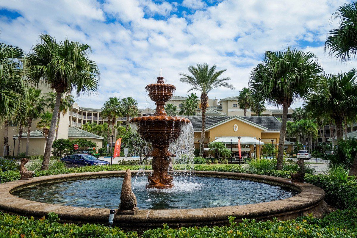 Ornate fountain in a round basin surrounded by lush greenery, buildings, and palm trees under a cloudy sky.