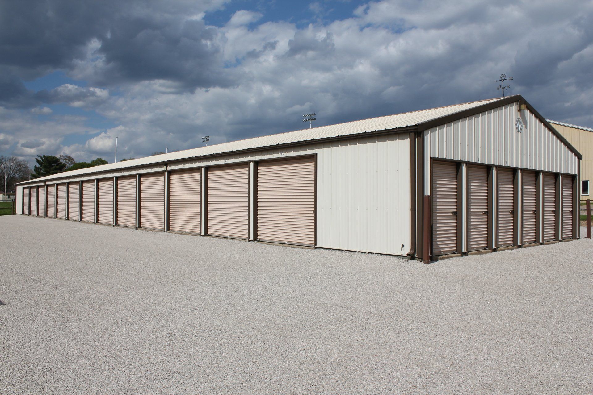 Storage units in a row, with tan doors, white building, gravel parking, and cloudy sky.