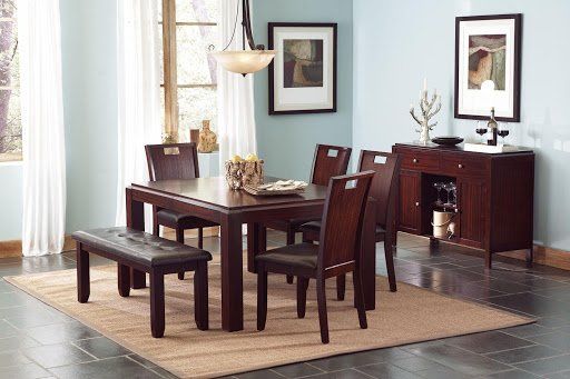 Dining room with a dark wooden table, chairs, and a bench, on a beige rug against a light blue wall.