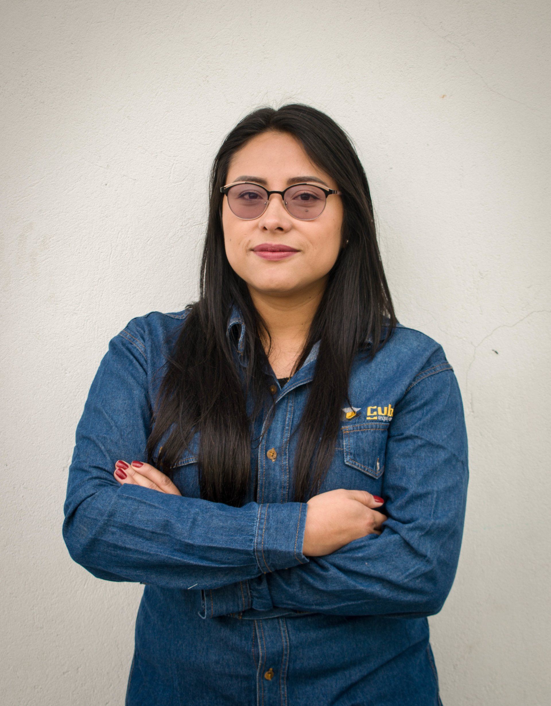 Una mujer que lleva gafas y una camisa vaquera está parada con los brazos cruzados.