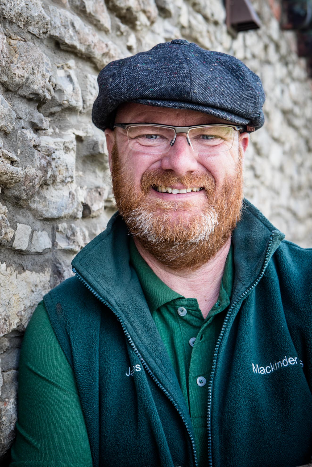 A man with a beard and glasses is wearing a hat and a green vest.
