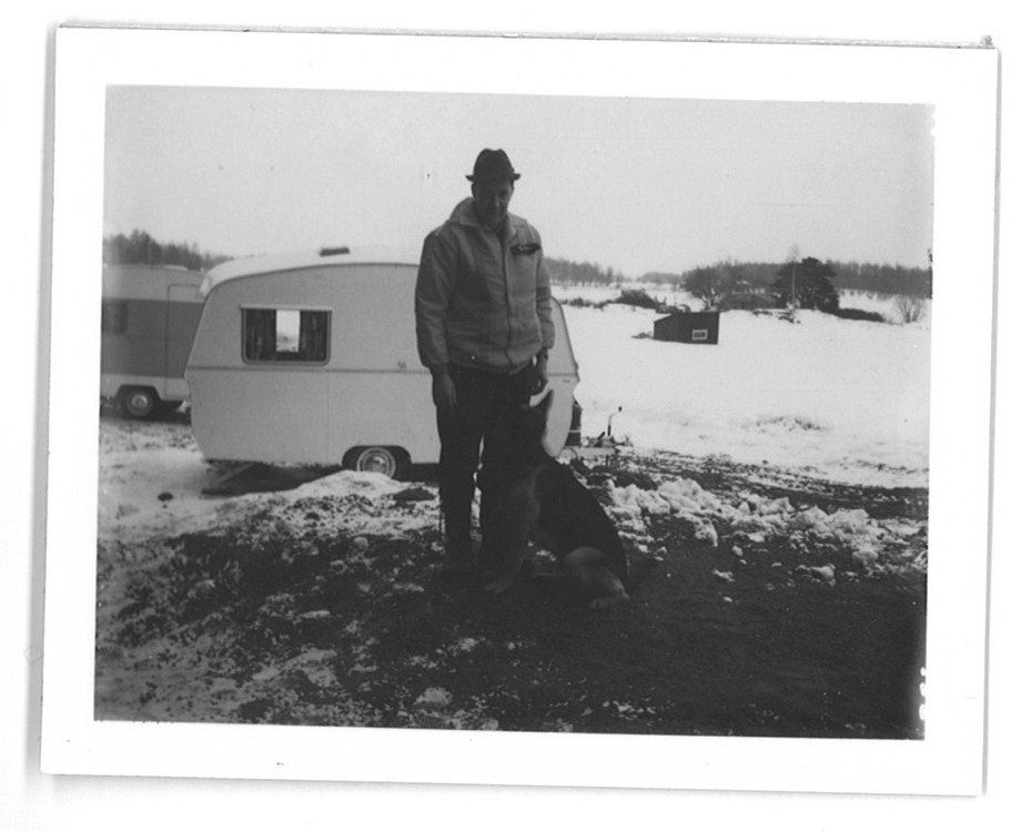 Man standing in the snow next to a small camper. He is wearing a jacket and hat. Winter scene.