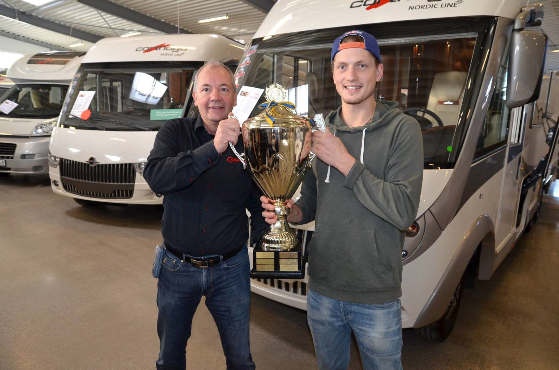 Two men holding a large trophy in front of RVs in a showroom. One man is smiling and the other looks serious.