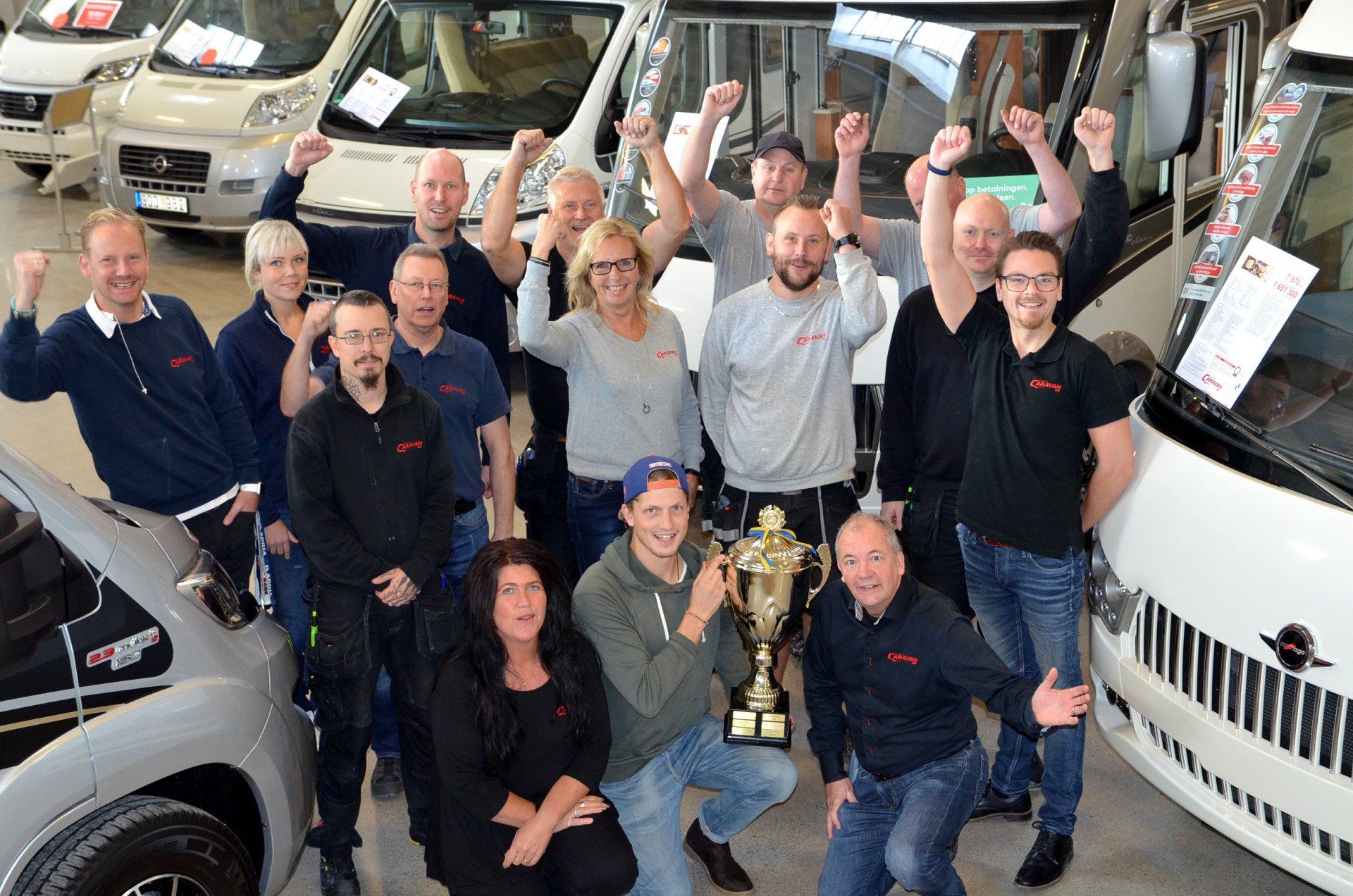 Group of people celebrating, holding a trophy in a car dealership, raising arms, smiling.