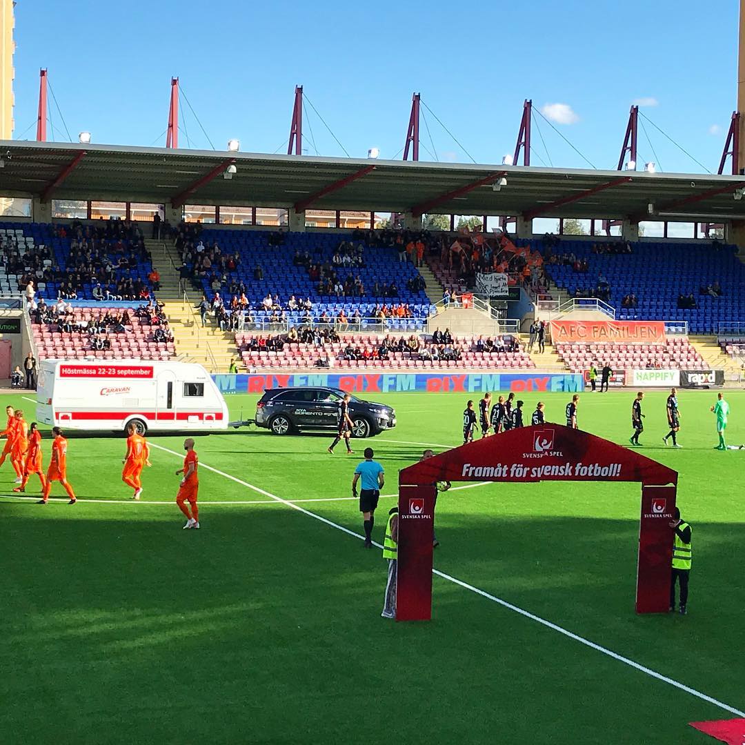 Soccer stadium with players entering the field under a banner, fans in stands, ambulance on sidelines.