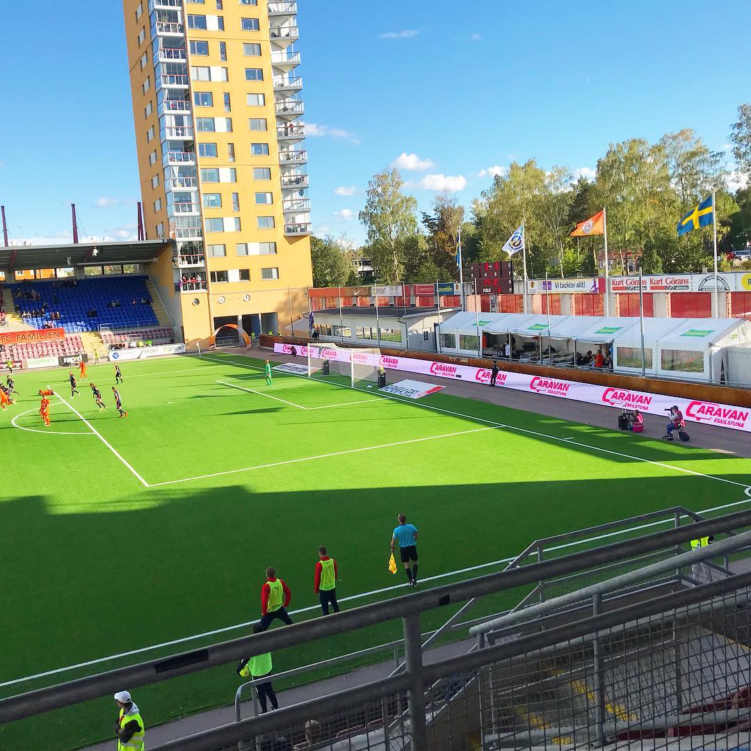 Soccer stadium with players on the field. Tall building in background. People in the stands. Blue sky.