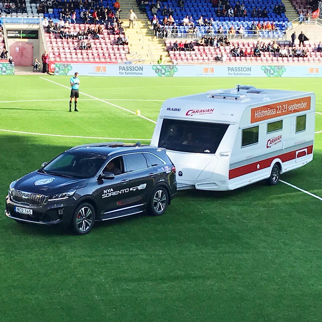 Black SUV towing a white camper on a green soccer field in front of a stadium crowd.