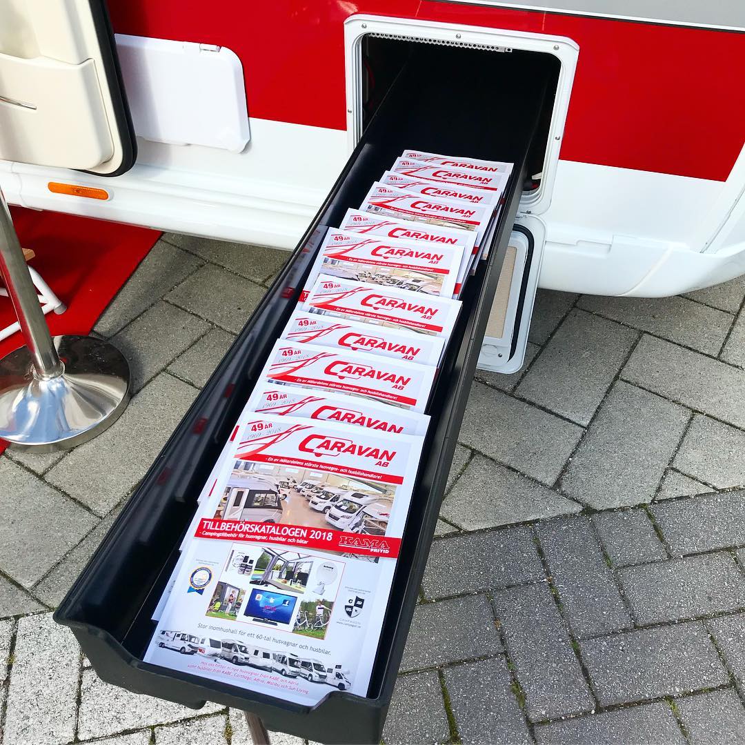 Open compartment of a caravan holding stacked brochures with red and white branding on a brick patio.