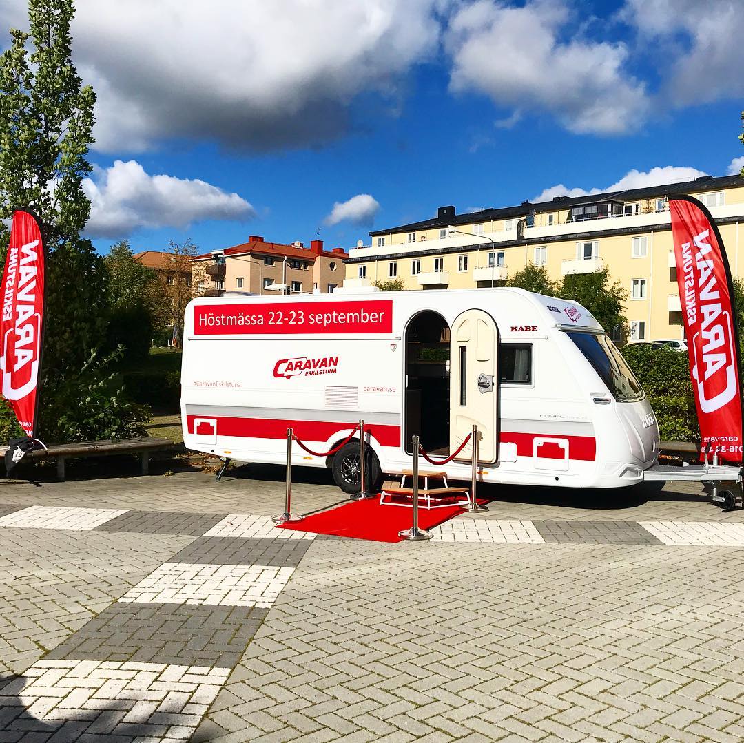 White and red caravan with red carpet and flags in a city square under a blue sky.