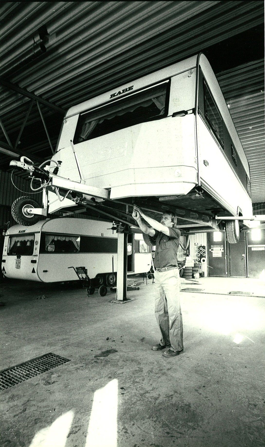 A person working on a raised white camper in a garage. Another camper is parked below.