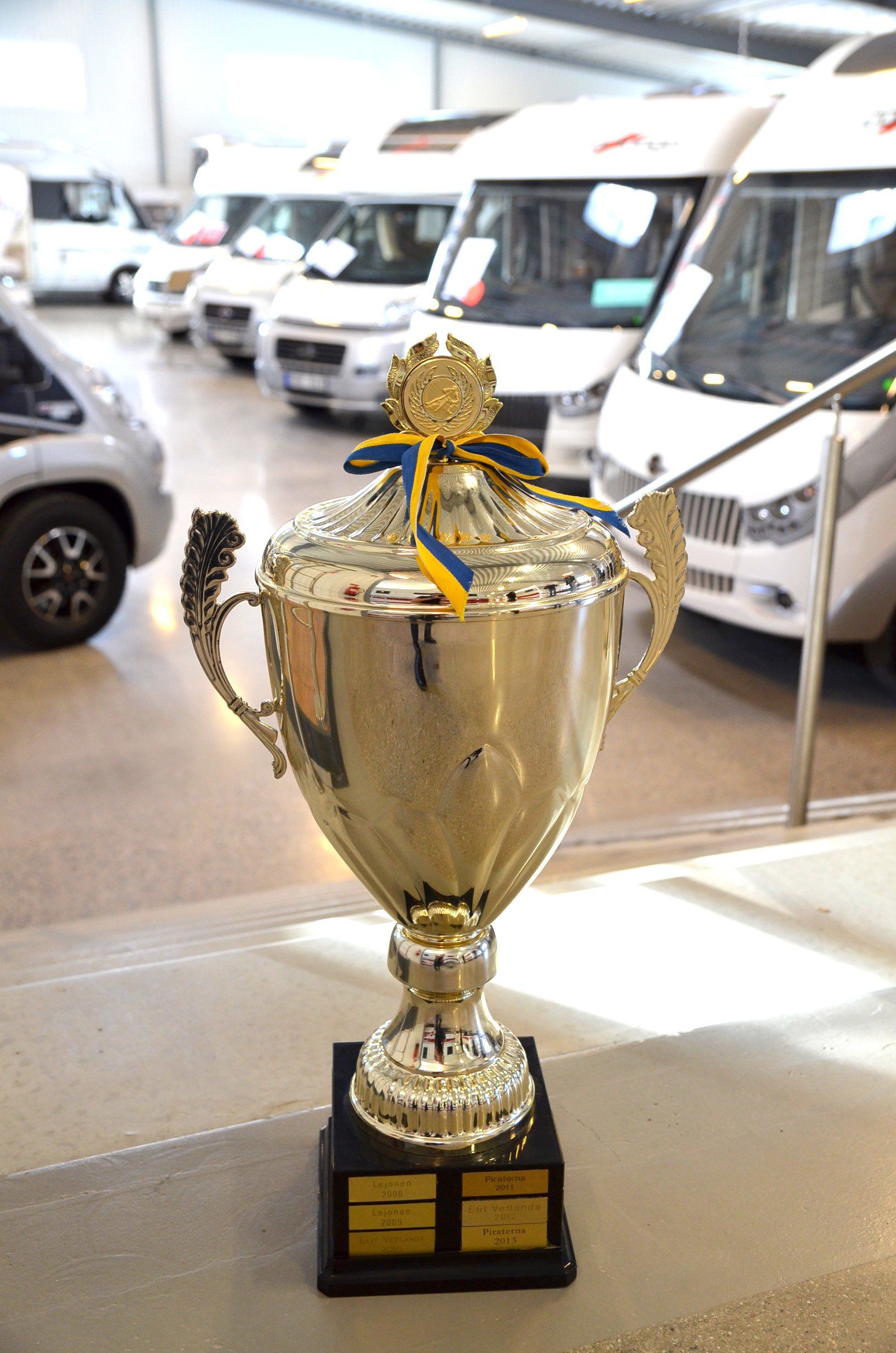 Large silver trophy on a black base, tied with blue and gold ribbons. RVs in background.