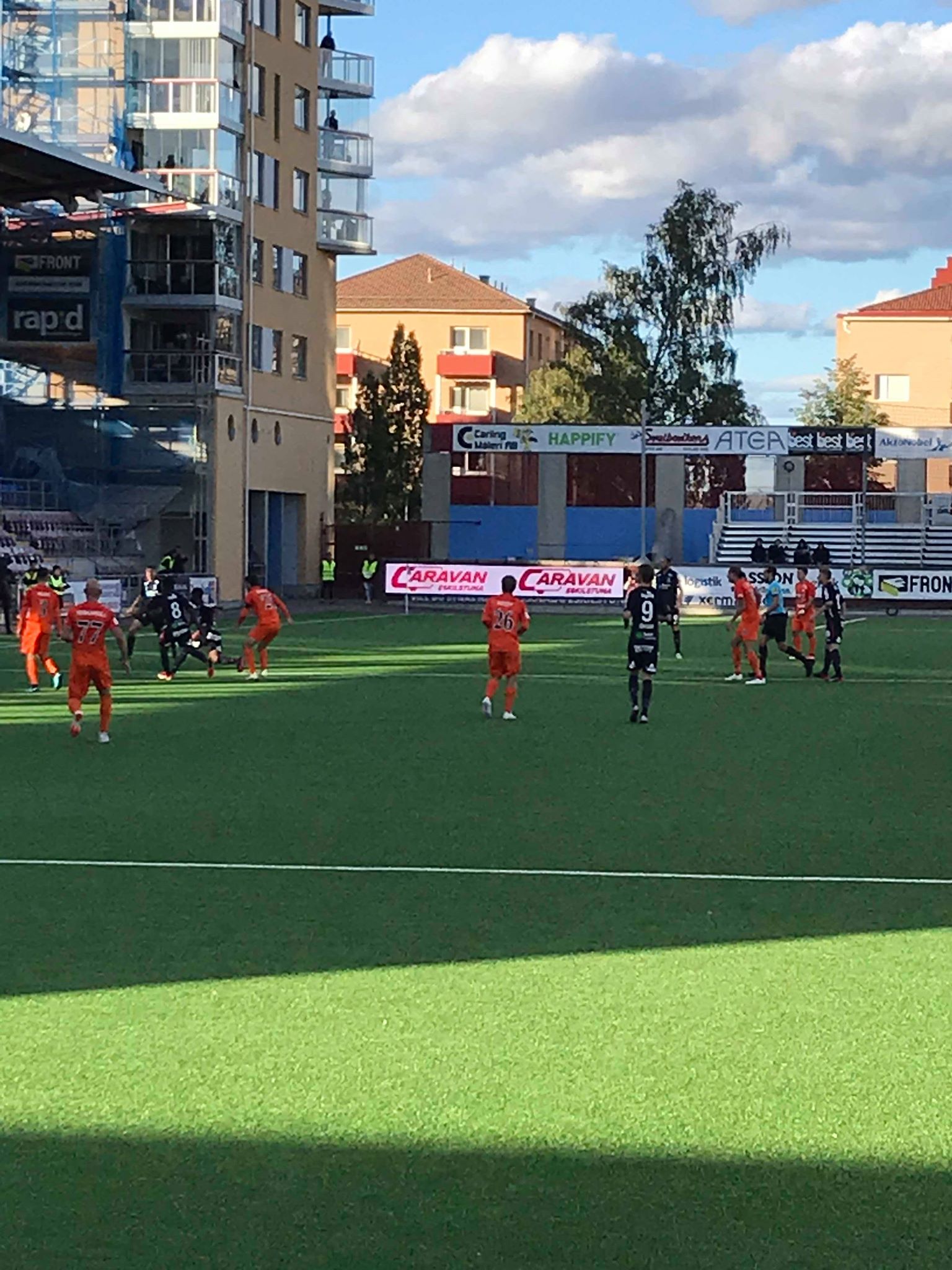 Soccer players in orange jerseys on a green field during a game, with buildings in the background.