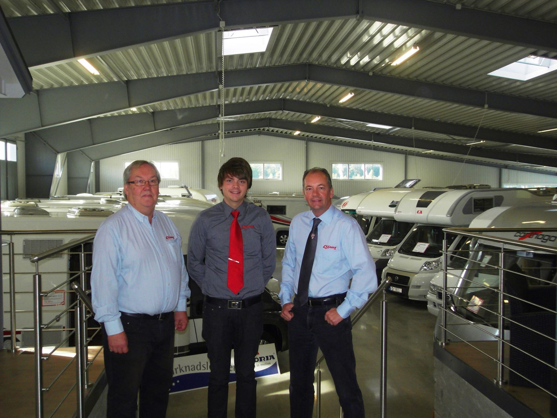 Three men in a showroom stand in front of recreational vehicles.