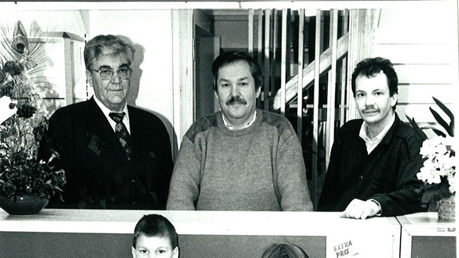Three men behind a counter, posing. One in a suit, two in casual sweaters. Interior setting.