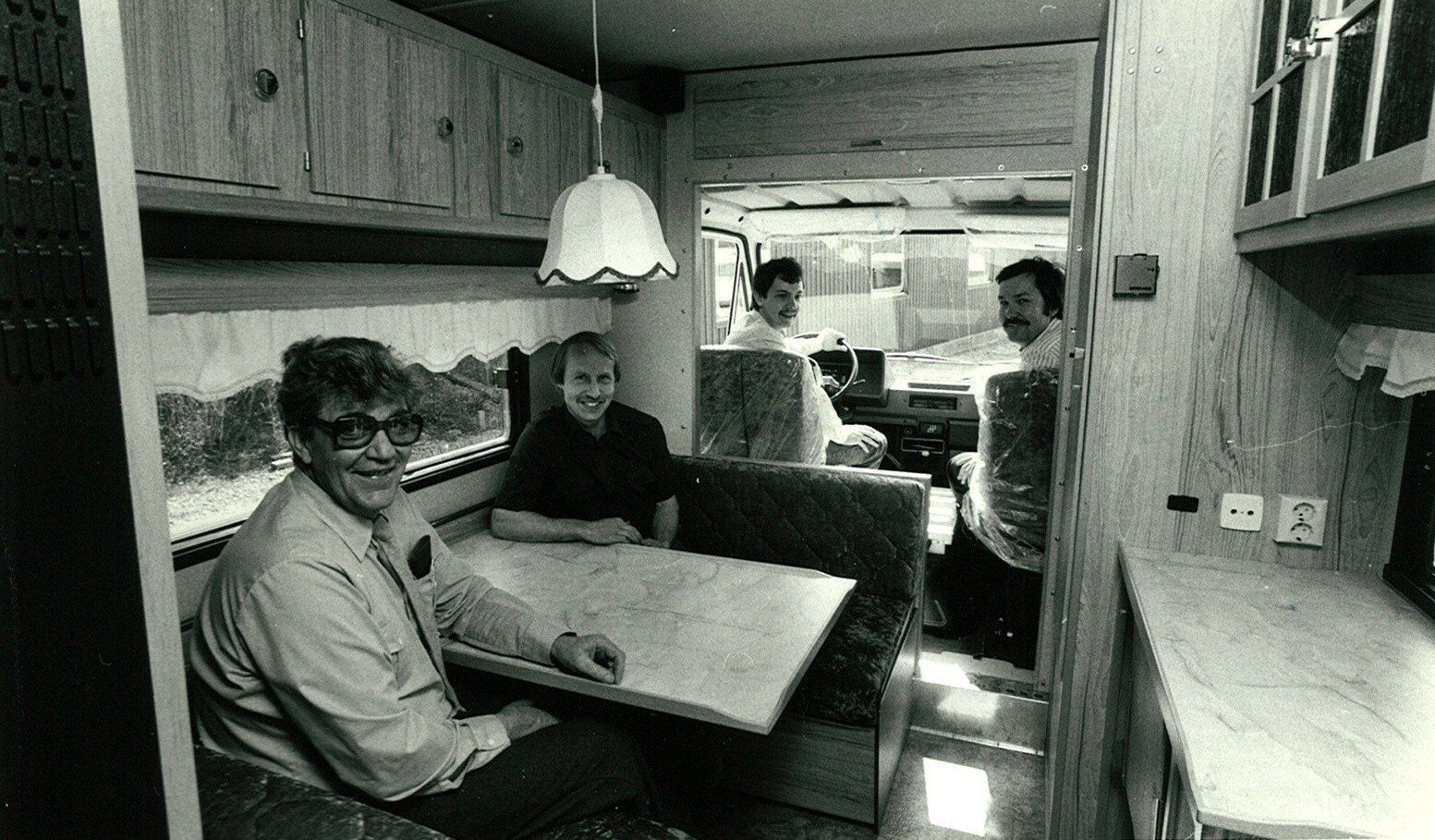 Interior of a recreational vehicle with four men; one sits at a table, three stand in the background.